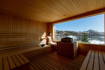 Young man relaxing in the sauna and watching winter forest through the window