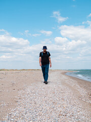 Smiling bearded millennial man in bucket hat black t-shirt walking on empty beach. Authentic male tourist lifestyle photo. Hipster guy outdoor. Solo travel adventure concept Active walking backpacking