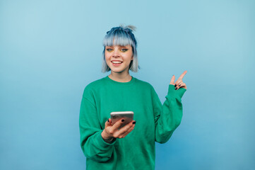 Happy teen girl with blue hair holds a smartphone and shows a finger to the side with a smile on his face isolated on a blue background.