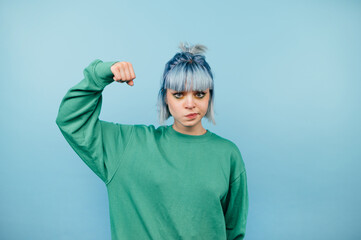 Serious teen girl in green sweatshirt stands on blue background and shows biceps to camera.