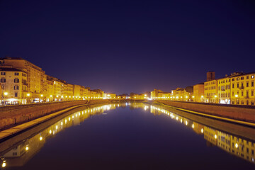 Fototapeta na wymiar Houses lit with golden light and their reflections in the Arno River at night in Pisa, Italy. Panoramic view