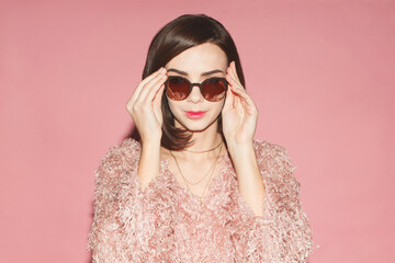young fashion woman adjusts sunglasses in pink dress isolated on pink background