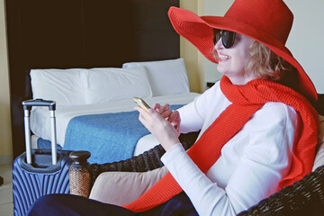 Woman wearing the big red hat with suitcase checking out of hotel with her baggage sitting on bed waiting for taxi ordered on mobile phone app,