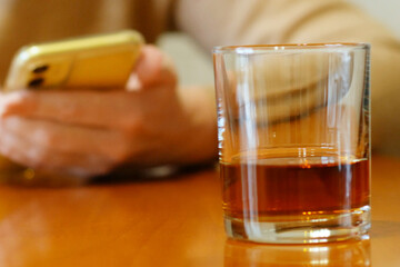 whiskey glass standing on a table on background of hand with mobile phone, hand alcohol order online concept