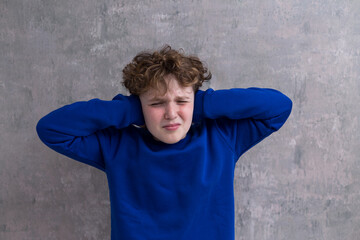 Horizontal shot of boy in blue sweatshirt cringing and holding his hands over his ears reacting to...