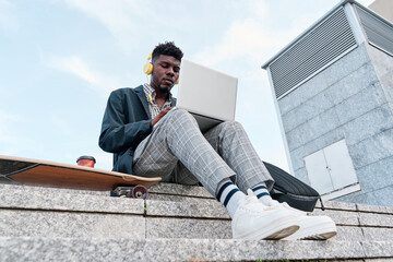 View from below of a young African-American adult man on a skateboard while working on a laptop...