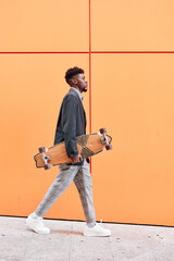 side view of a young man with blazer and skateboard walking down the street against an orange...