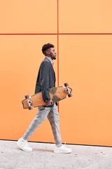 Poster side view of a young man with blazer and skateboard walking down the street against an orange background. © Oscar