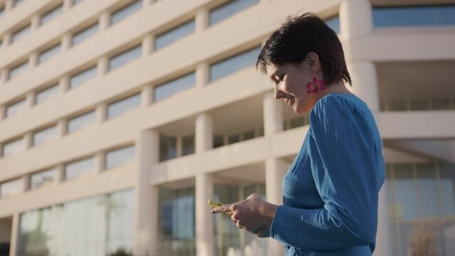 Attractive young girl with short haircut, stylish earrings in blue romantic dress during weekend, travel, vacation walking on street city center. Pretty blogger chatting on smartphone, smiling