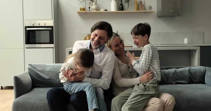 Tickling and giggling. Joyful family couple with two children siblings having fun playing laughing on comfy couch at new modern studio apartment. Excited kids enjoy funny activity with beloved mom dad