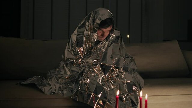 Mid shot of a middle-aged man sitting in a dark room, wrapped in a large shiny material. Man suffers from electromagnetic hypersensitivity and hides under a metal blanket in dark room with candles. 