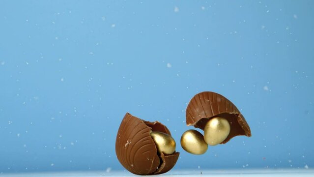 Animation of chocolate easter egg falling and breaking open with gold eggs, on blue background