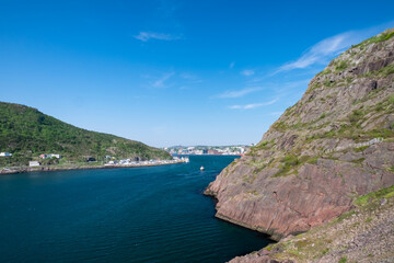A footpath, Signal Hill hiking trail or path along a hillside. The cliff is rocky with grass patches. The city of St. John's, Newfoundland, is in the background on a sunny day. The sky is bright blue.