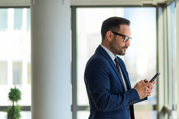 Fototapeta premium Young businessman standing next to window holding phone