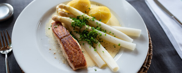 White asparagus with hollandaise sauce and fried salmon on a white plate in a restaautant. Close-up. Seasonal gastronomy presented in a modern way. Top view.  - 498133284