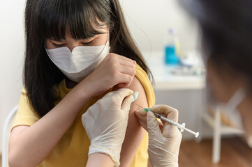 Asian woman nurse injecting covid-19 vaccine to young Thai girl patient wearing face mask in clinic...