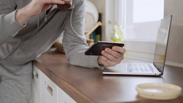 unrecognizable man wearing gray hoodie holding smartphone watching video or playing game drinking coffee in the kitchen