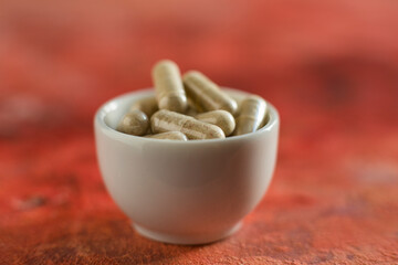 herbal medicine, capsule pill with herb in white bowl on red background for alternative medical concept