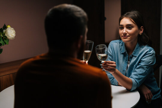 Rear View Of Happy Young Couple Clinking Glasses Of White Wine Sitting At Table With Candles In Dark Cozy Room. Loving Man And Woman Celebrating Anniversary, Valentines Day, Enjoying Romantic Date.