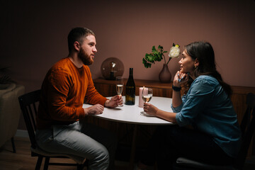Side view of happy young couple enjoying talking and sitting together at dinner table with candles in cozy dark room. Love couple celebrating anniversary or Valentines day having romantic dinner