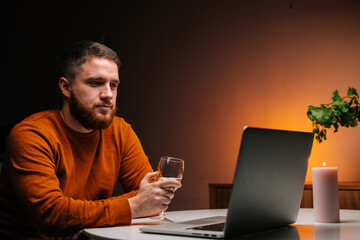 Handsome young man enjoying distant dating using laptop computer, holding glass with wine, talking celebration toasting via video call looking on screen. Couple in love chatting on remote date.