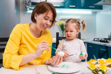 Easter celebration and traditions. Smiling mother and her toddler daughter painting eggs. Young woman teaching a little girl paints egg