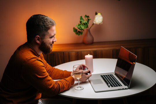 Cheerful young man making video call using laptop computer presenting ring and making remote marriage proposal sitting at table. Loving boyfriend making distant proposal for marriage, marry me.