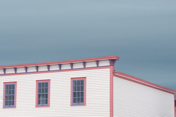 The roof section of a large white vintage wooden building with decorative pink and purple wood trim. There are three multi-pane windows on the top floor. The background is a dramatic blue cloudy sky.