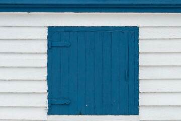 Fototapeta na wymiar A vibrant royal blue wooden shutter door on a white wood wall. The exterior of the building is covered in white horizontal beveled clapboard siding. The boards on the window are vertical with hinges. 