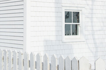 The exterior corner of a vintage wooden building with a closed glass single hung window. The wall is covered in white shingles and a shadow. There's a white picket fence attached to the building. 