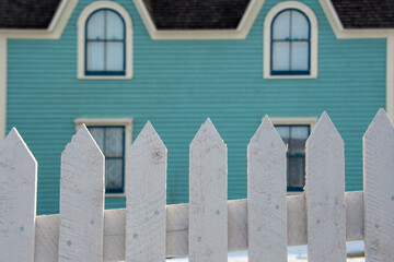 Multiple windows on a blue exterior wall of a vintage style building. The house has narrow clapboard on the wall. There's a white picket fence in the foreground with a mound of white snow. 