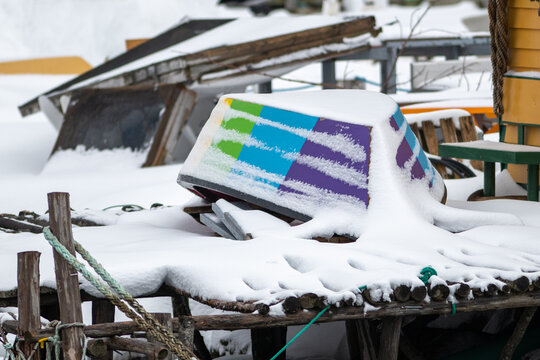 A wooden traditional fishing boat turned bottom up on a wharf. There's fresh white snow on the small boat. The vessel is painted purple, blue, green, yellow, and red as in the gay pride flag.  