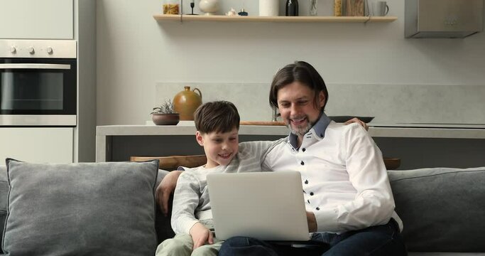 Happy foster dad adopted son teenager cuddle on couch focused on laptop screen watch video read sport news play game. Caring father software engineer teach tween boy to make simple program code on pc