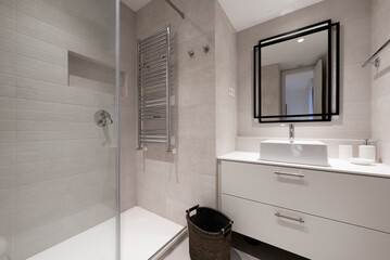 Bathroom with mirror with black metal frame, white porcelain sink, gloss white dresser, marble tiles, white fixtures and chrome metal hanger inside shower stall with glass partition and wall niche