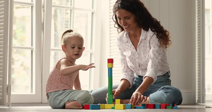 Young Hispanic nanny sit on floor with underheating play creative game with little preschool girl help to build from blocks. Mom and small child daughter construct high tower from colorful brick set