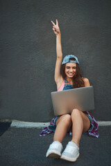 Getting work done while chilling hard. Shot of a cheerful young woman working on a laptop while raising her hand and being seated against a grey background.