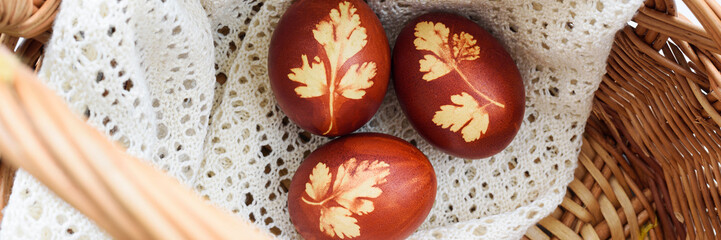 Panoramic photo. Top view of easter eggs, lying in wicker basket, on a lace doily. Painted brown, with a plant theme. Easter basket.