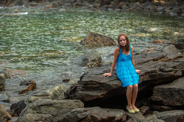 A teenage girl in a blue dress sits on a rocky sea beach.