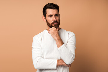 Fototapeta premium Thoughtful brunette man with beard in white t-shirt holding his chin and pondering