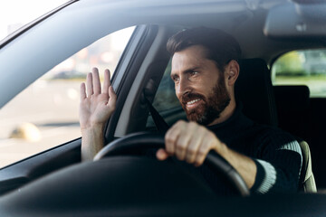 Handsome young smiling driver of car waving hand as a sign of greeting while driving the car