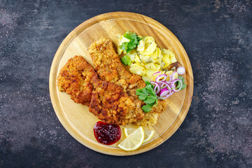 Traditional deep-fried schnitzel with potato salad, cranberry jam and lemon slices served as top view on a rustic wooden board with copy space