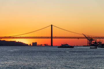 Sunset with orange sky behind the 25 de Abril Bridge, 25th of April Bridge, a red suspension bridge over the Tagus river, Lisbon, Portugal