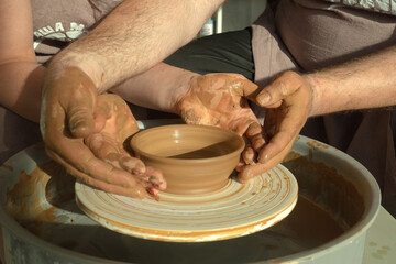 Warm closeup of pottery hands of a man and a woman shaping clay on pottery wheel. Making ceramics vase in sunlit workshop. Dating concept