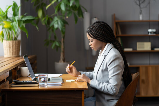 Focused African Female Office Assistant With Braided Hairstyle Writing Something Down On Sticky Note While Sitting In Front Of Laptop, Black Woman Freelancer Planning Out Day While Working Remotely