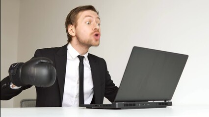 Young angry businessman in black suit wearing boxing gloves using laptop. - Powered by Adobe