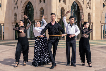 A flamenco dancer dancing with a cane next to 4 other dancers posing in front of a building with...