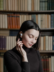 Woman in wireless headphones listening audiobooks on background of library shelves with paper books