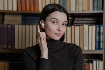Woman in wireless headphones listening audiobooks on background of library shelves with paper books