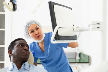 African-american man sitting on dental chair. Asian woman dentist pointing at display.