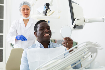 African-american man looking at his teeth through mirror after dental restoration procedure. Asian woman dentist standing in background.